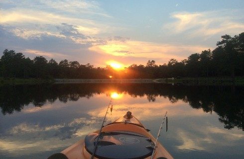 A lone orange kayak with fishing poles out on a calm body of water surrounded by trees at sunset
