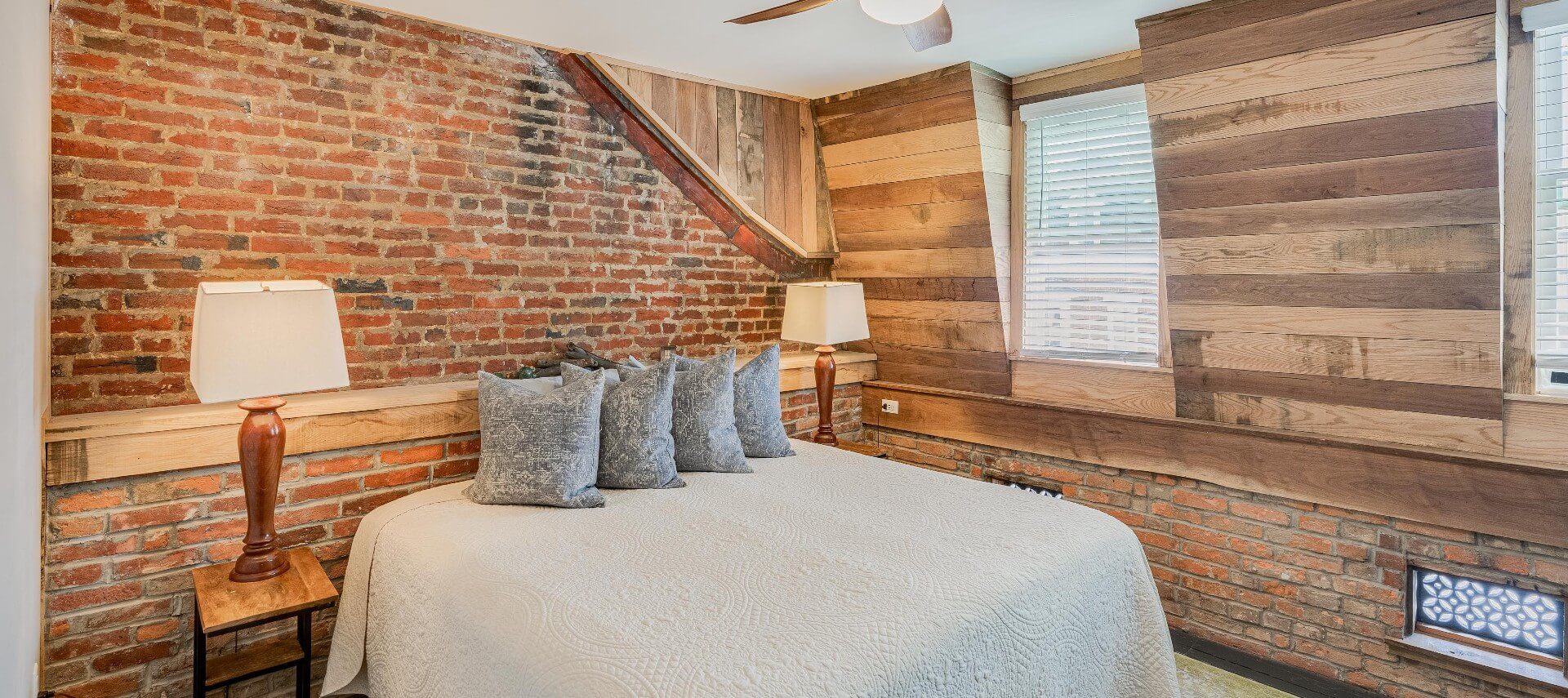 Unique bedroom with king bed, brick and wood panel details on the walls and two bright windows