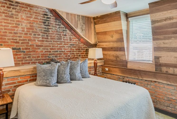 Unique bedroom with king bed, brick and wood panel details on the walls and two bright windows